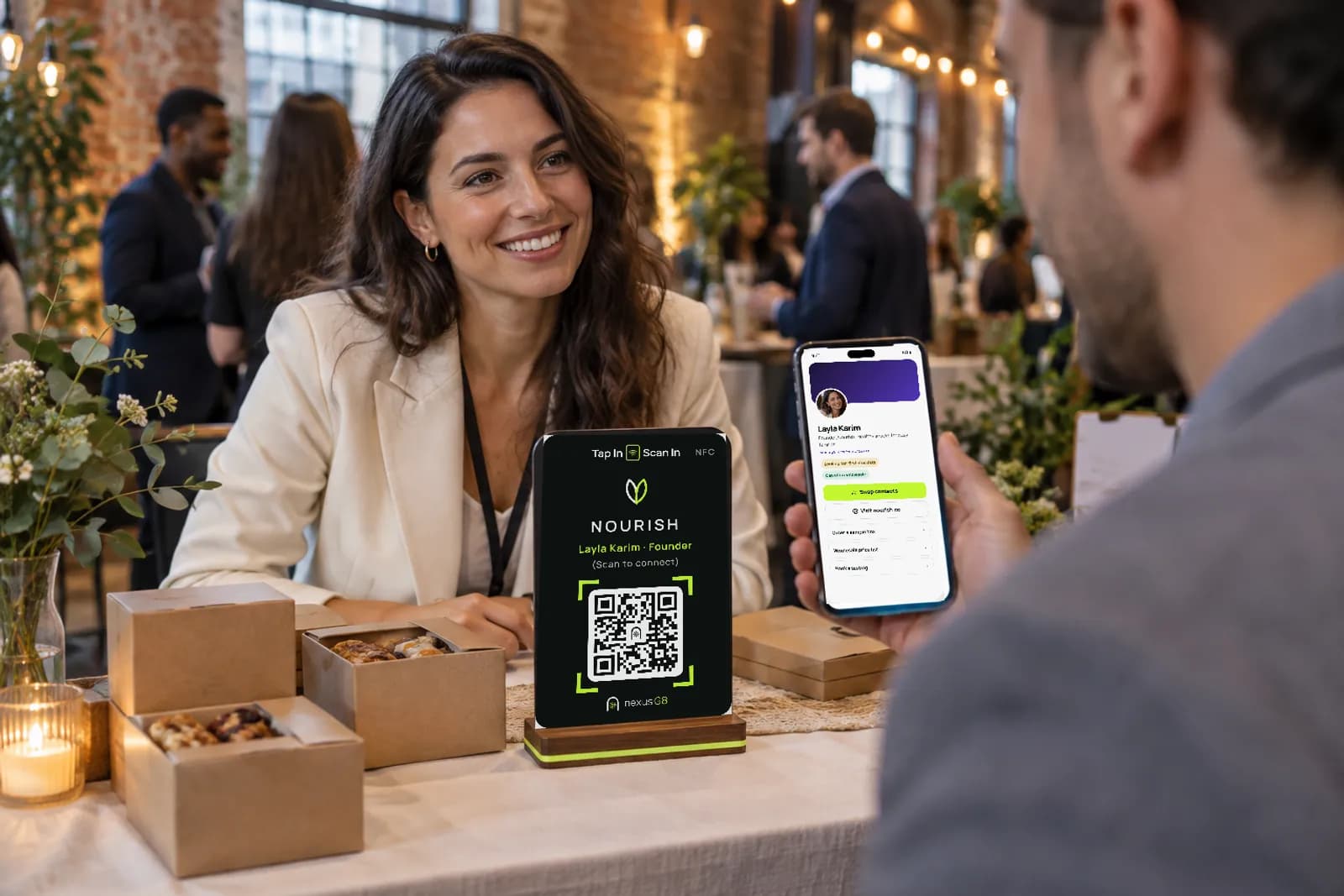 At a startup showcase a founder smiles across her table, where her Nourish table-top gate stands, while a visitor holds up his phone showing her NexusG8 page with Swap contacts and Visit website